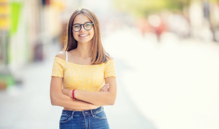 young girl with glasses standing in somewhere the city smiling and looking at the camera.